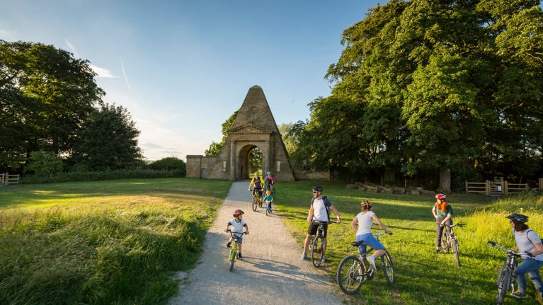 Families and cyclists ride along a sunlit path leading to the Obelisk Lodge at Nostell, surrounded by green trees and open grassland.